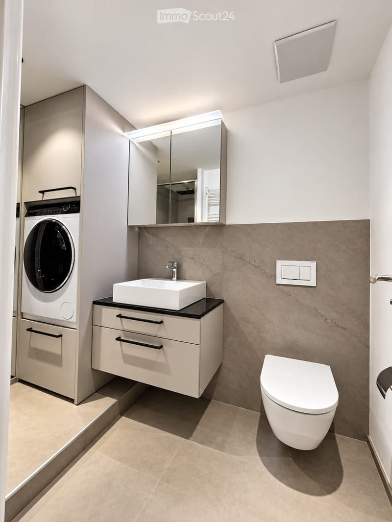 Modern bathroom with a white sink, mirror, toilet, and a built-in washing machine. Features a sleek, contemporary design with light-colored tiles and a minimalist aesthetic.