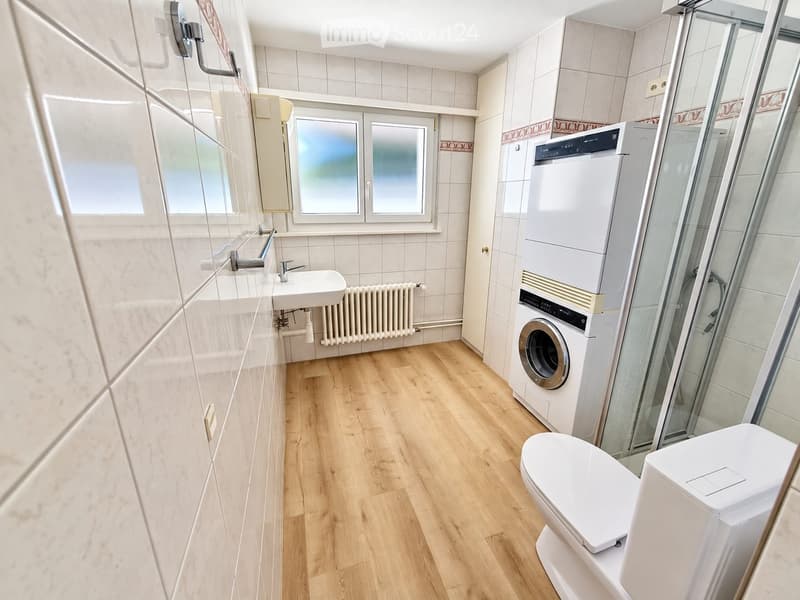 Modern bathroom with wooden flooring, white tiles, a sink with a faucet, a toilet, a washing machine, a mirror, and a radiator.