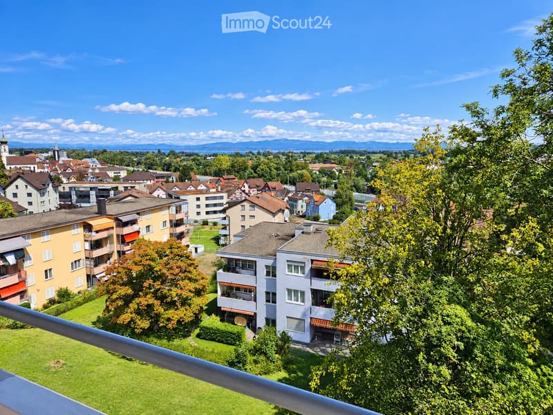 A panoramic view of a residential area with multiple apartment buildings, trees, and a distant view of a town and mountains under a clear blue sky.