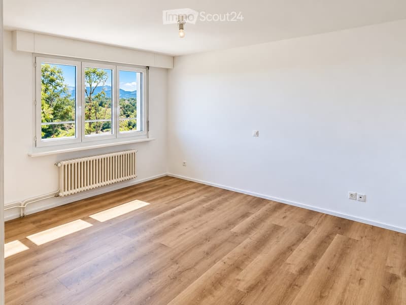 Empty room with light wooden flooring, white walls, large windows with scenic view, radiator, and electrical outlets.