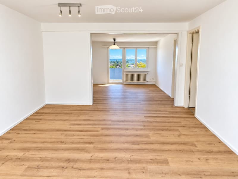 Empty hallway with wooden flooring, white walls, and a view of a room with large windows and a radiator.