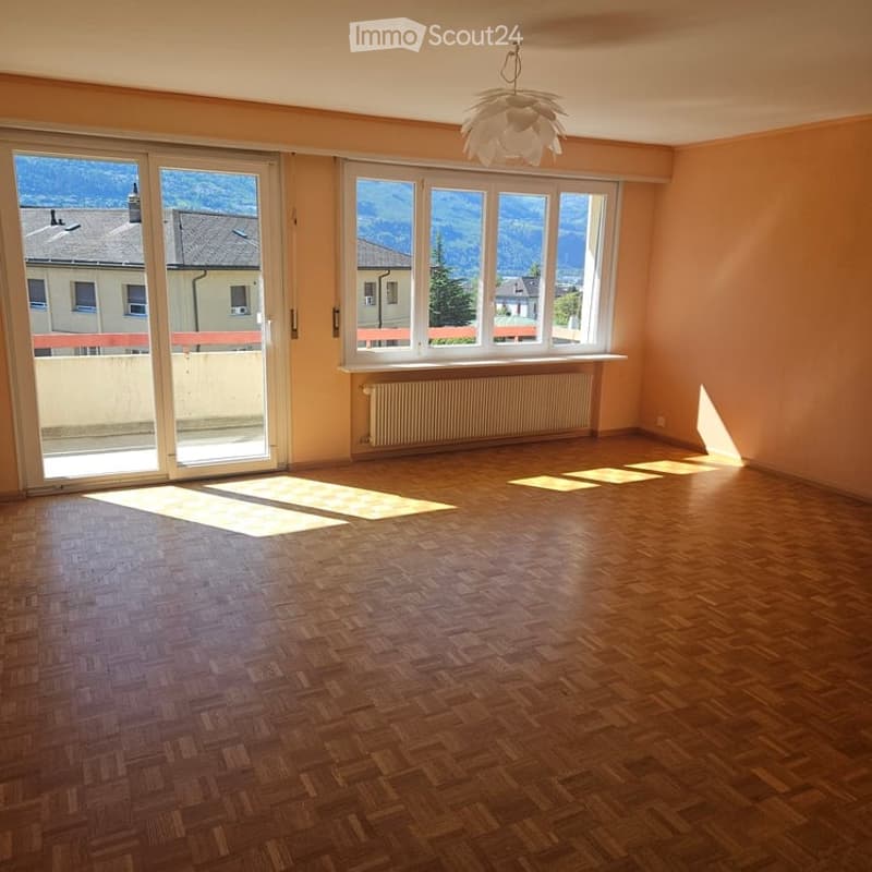 Empty room with wooden flooring, large windows offering a scenic mountain view, and a hanging light fixture.