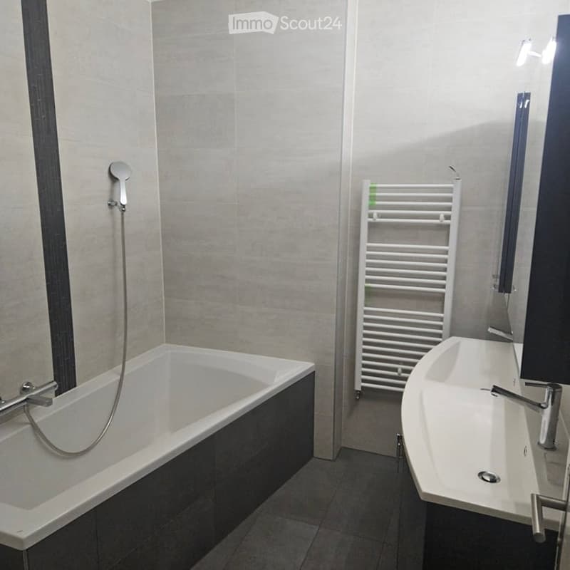 Modern bathroom with a bathtub, sink, and a white radiator. The walls are tiled in light grey, and the floor is dark grey tiles.
