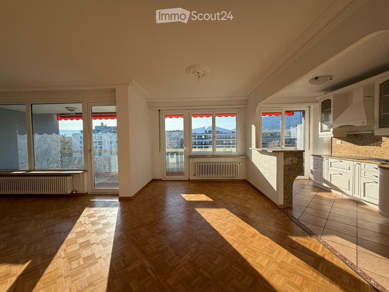 Bright, unfurnished room with wooden flooring, large windows offering a scenic view, and a small kitchen area with cabinets and a countertop.