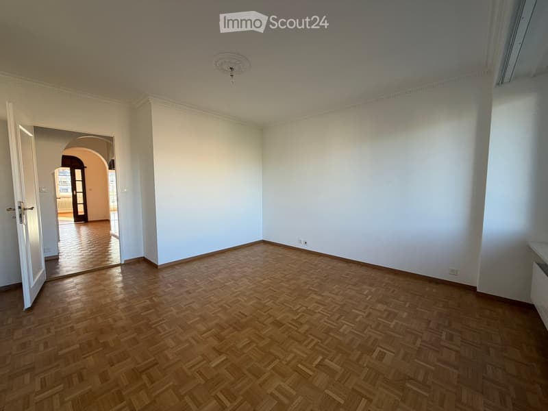Empty room with wooden herringbone flooring, white walls, and an open doorway leading to another room.