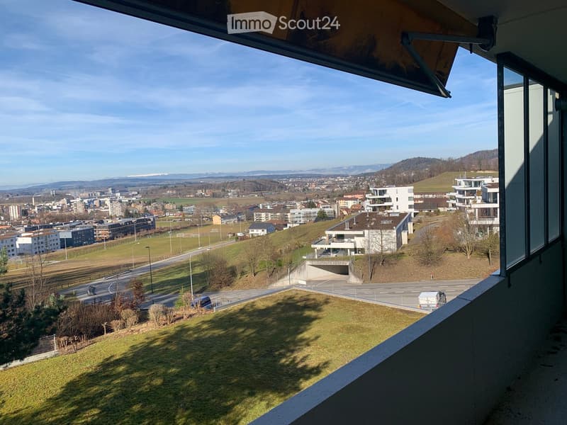 Balcony overlooking a suburban area with buildings, roads, and distant hills under a clear sky.