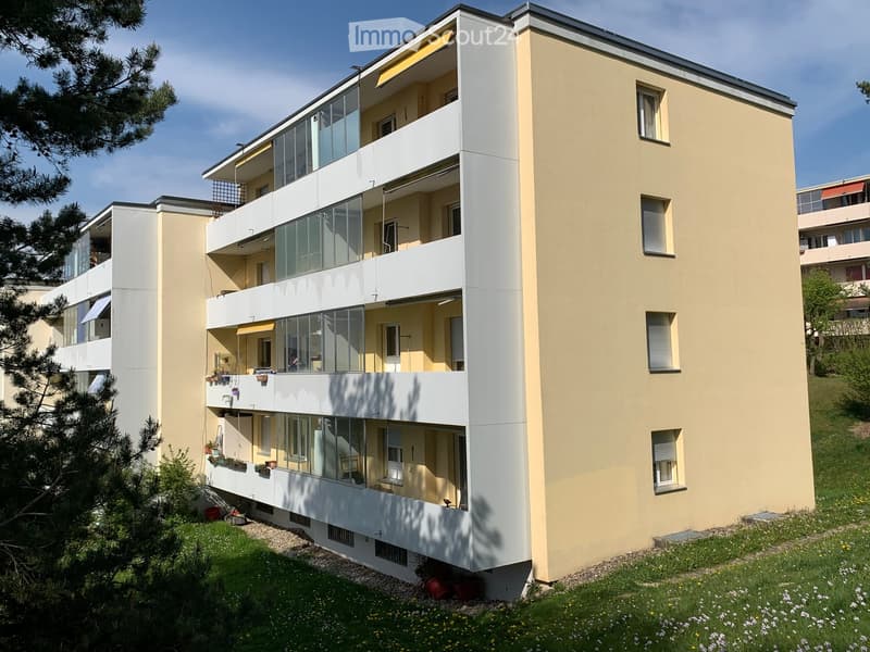 Modern multi-story residential building with beige facade, large glass windows, and balconies. The building has multiple floors with visible windows and railings, surrounded by greenery and a grassy area.