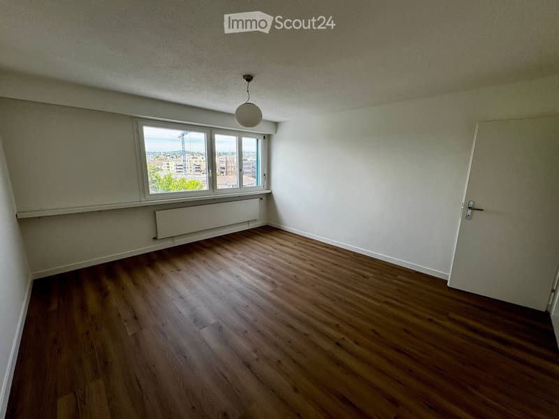 Empty room with wooden flooring, white walls, a window with a view, a radiator, and a single light fixture on the ceiling.