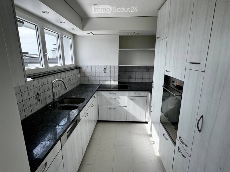 Modern kitchen with white cabinets, black countertop, double sink, and built-in oven. Features large windows and tiled backsplash.