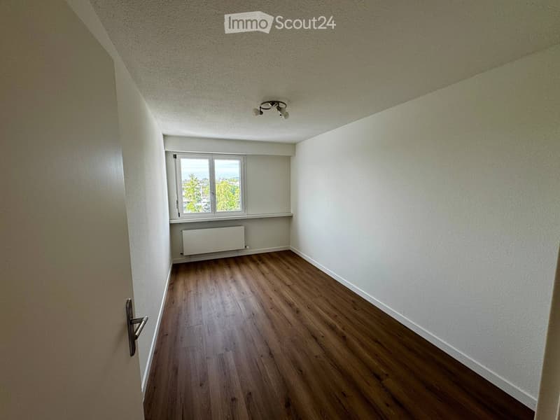 Empty room with wooden flooring, white walls, and a window with a view of greenery. Features a radiator and a ceiling light fixture.