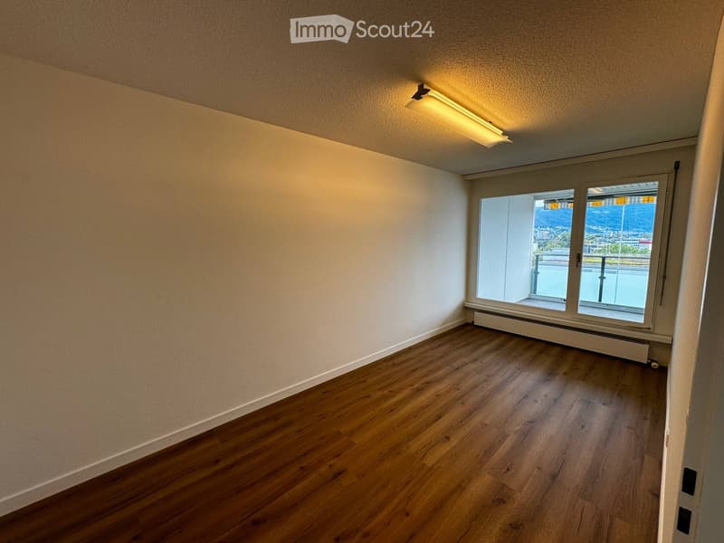 Empty room with wooden flooring, white walls, and large windows offering a view of the city and mountains.