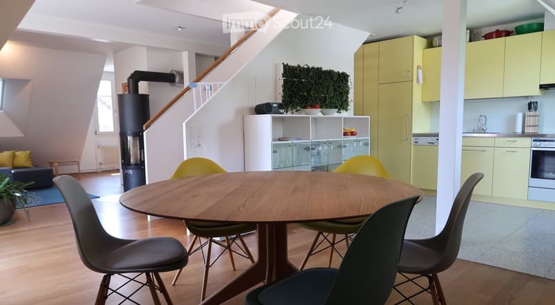 Open-plan living and dining area with a round wooden table, chairs, sofa, kitchen area with cabinets, fridge, and oven, and a staircase leading to an upper level.