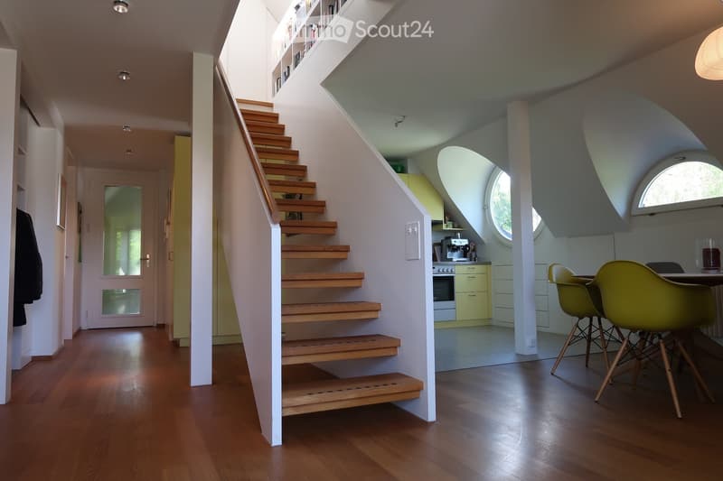 Modern hallway with wooden flooring, staircase leading upwards, built-in shelving, and a small kitchen area visible to the right.