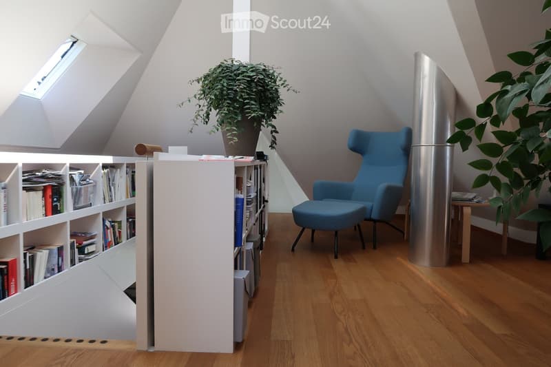 Modern, well-lit room with wooden flooring, white walls, and skylight windows. Features a white bookshelf filled with books, a blue armchair with matching stool, a small wooden table, and two large potted plants.