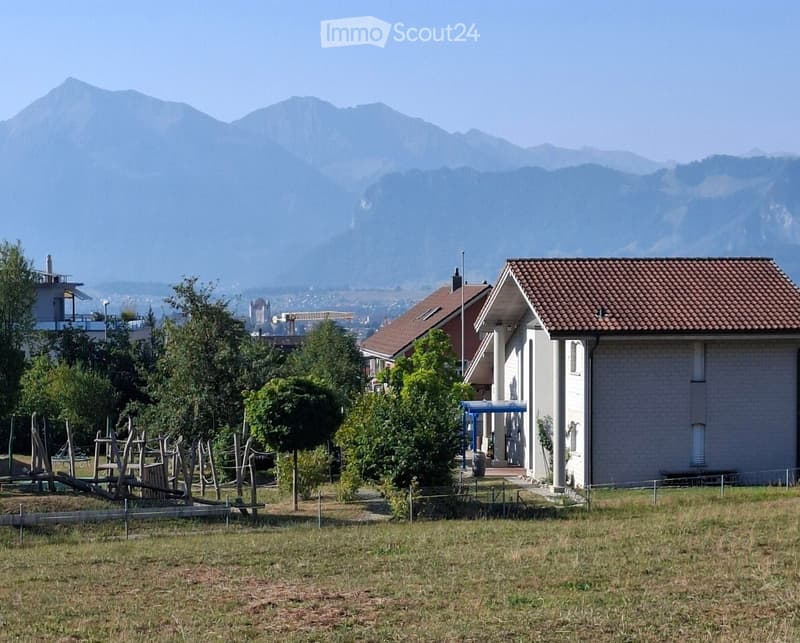A two-story house with a tiled roof and white exterior, surrounded by trees and a playground area, with mountains in the background.