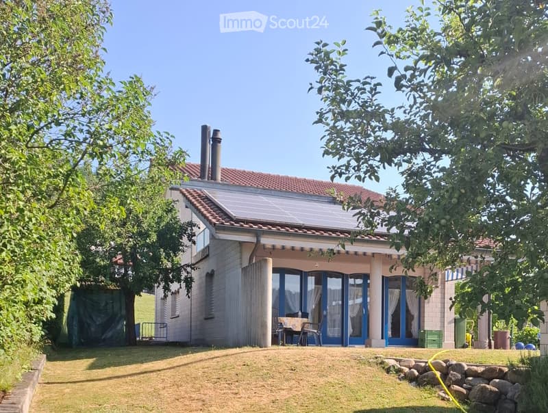 Two-story house with a red-tiled roof, solar panels, large windows, and a covered patio area with outdoor furniture.