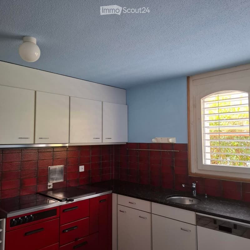 Modern kitchen with white cabinets, red lower cabinets, black countertop, sink, and oven. Features a window with blinds and a round ceiling light.