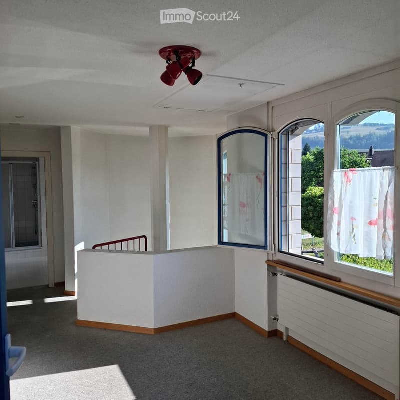 Empty room with white walls, carpeted floor, large windows with floral curtains, and a view of greenery outside.