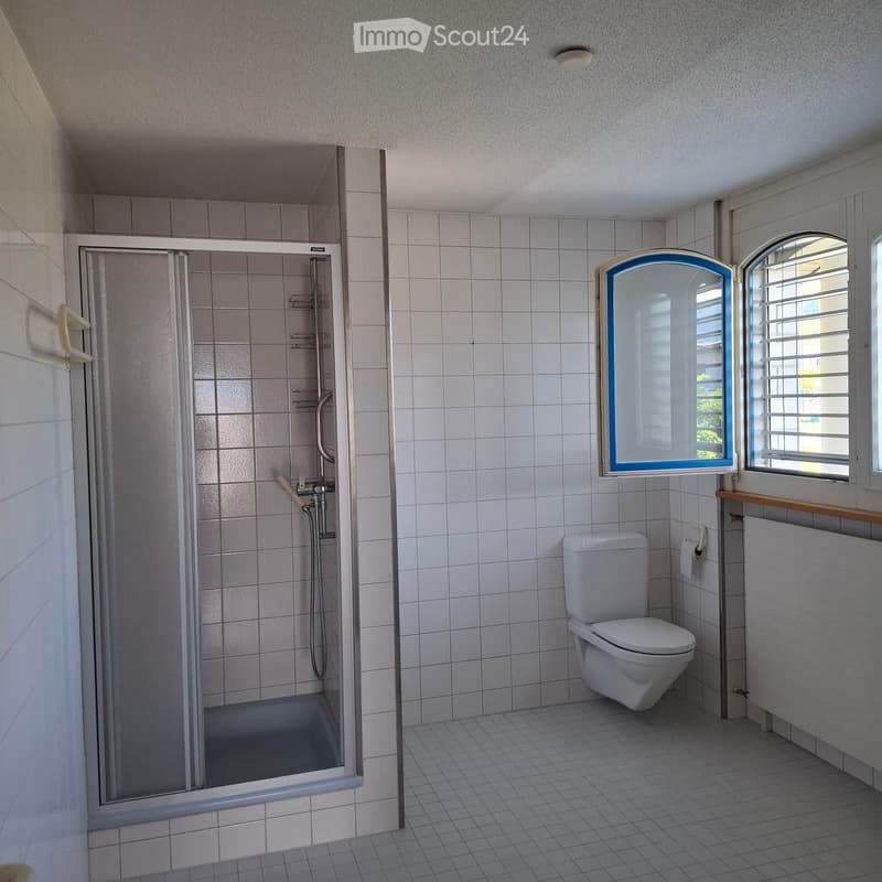 Clean bathroom with white tiled walls and floor, featuring a toilet, a glass-enclosed shower, and a window with blinds.