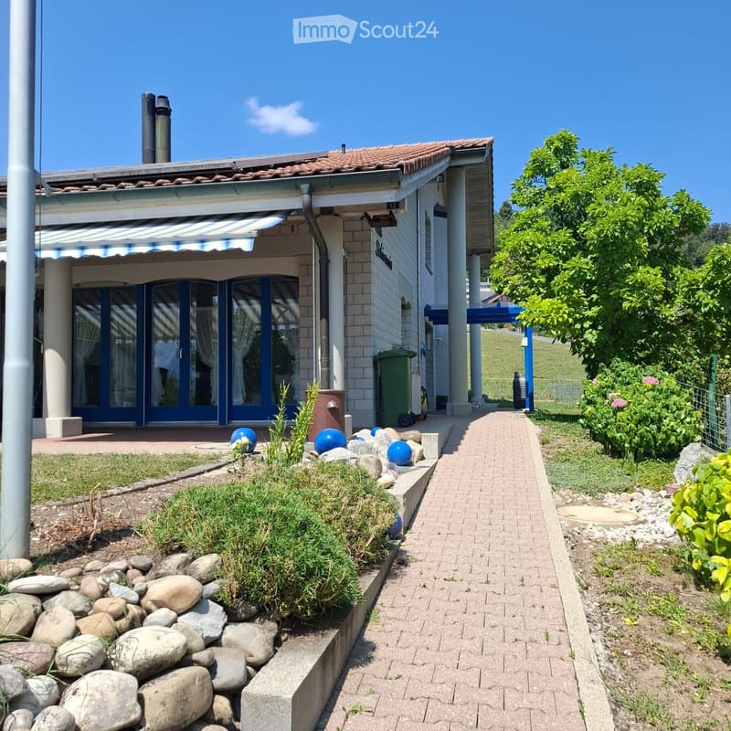 A well-maintained garden area with a brick pathway leading to a house entrance, featuring decorative rocks, plants, and a blue awning over the entrance.