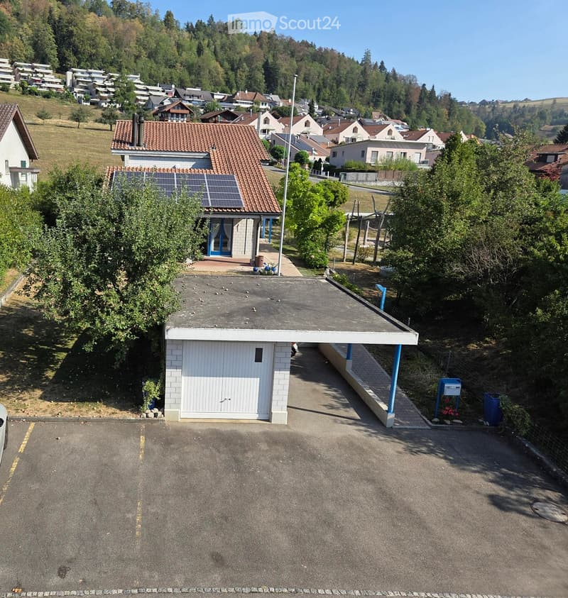 A two-story house with a red-tiled roof, solar panels on the roof, and a white garage. The property includes a paved driveway and a small parking area.