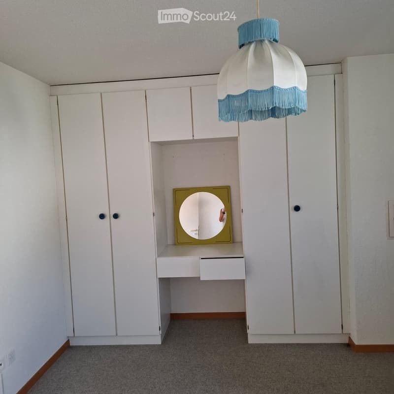 Empty room with white built-in cabinets, a small desk with a mirror, and a hanging light fixture with a white and blue shade.