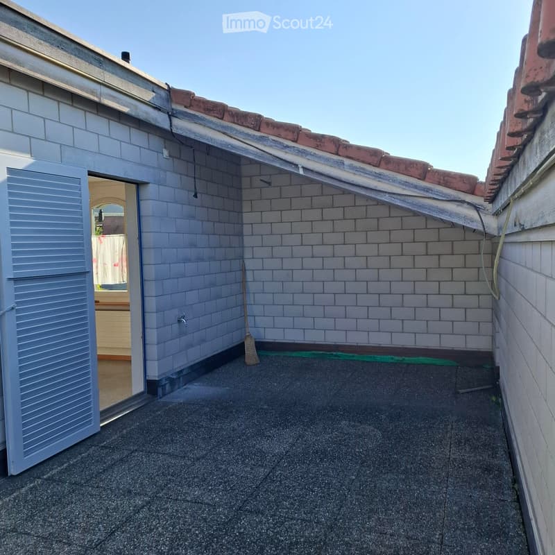 Small, enclosed rooftop terrace with grey brick walls, a concrete floor, and a broom leaning against the wall. An open door leads to an interior space.