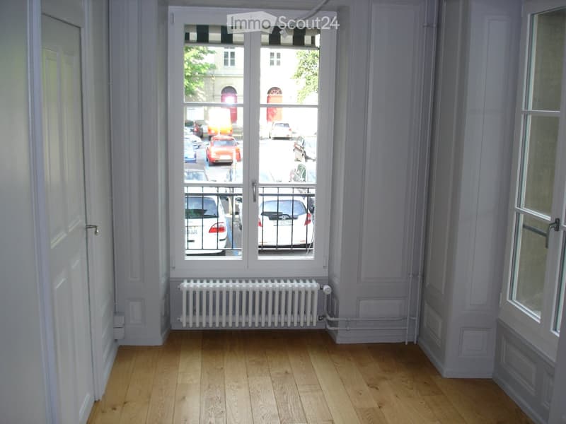Empty room with wooden flooring, white walls, a radiator, and large windows overlooking a street with parked cars.