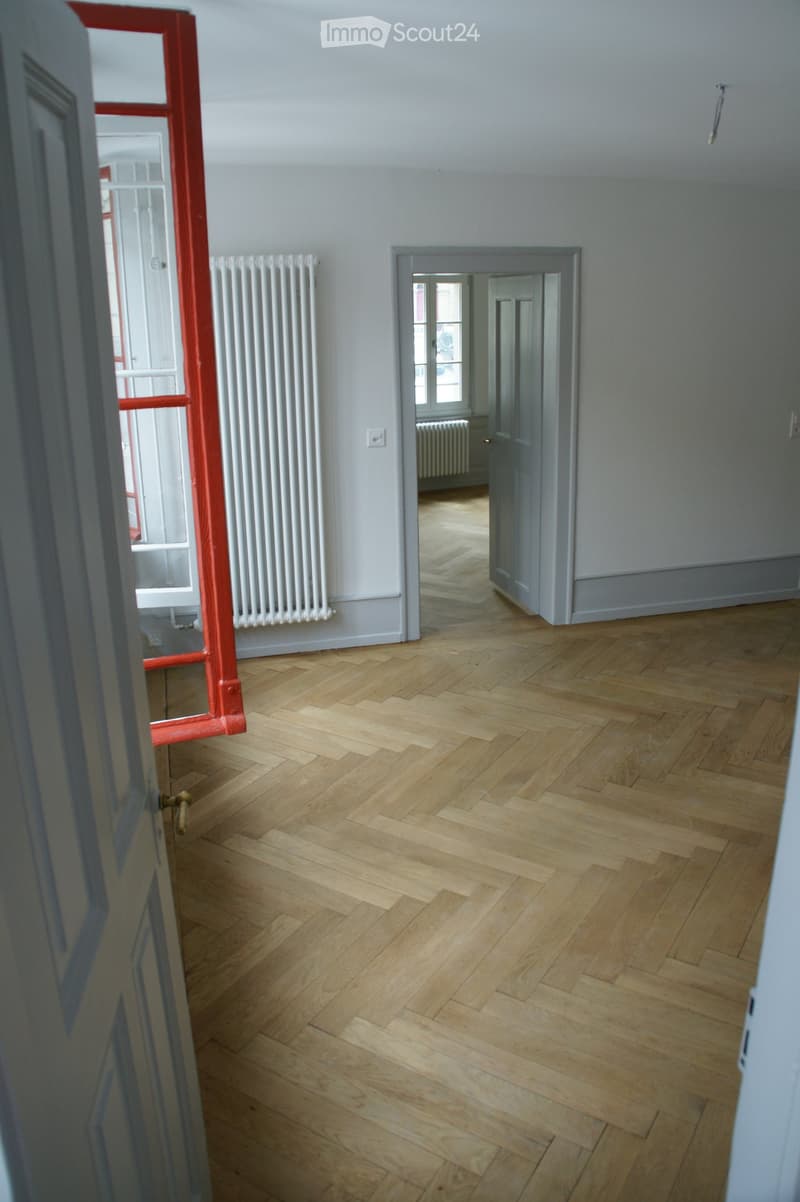 Empty room with wooden herringbone flooring, white walls, and a radiator. A doorway leads to another room with similar flooring.