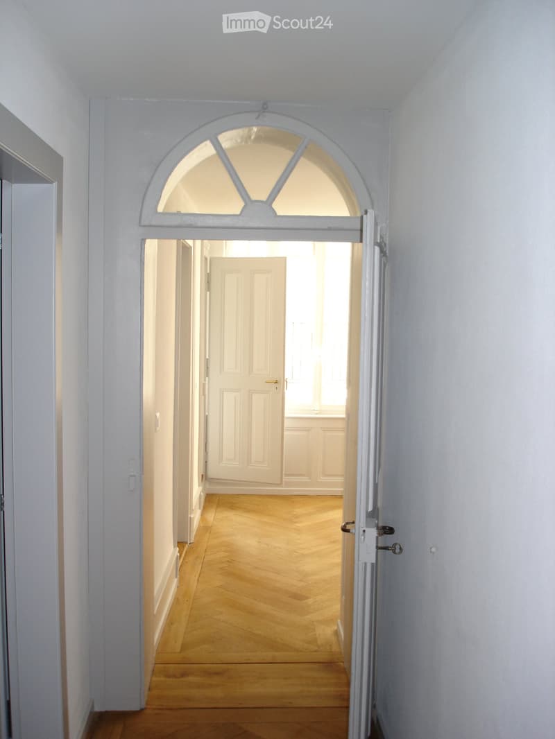 Empty hallway with wooden flooring, white walls, and a semi-circular arched doorway leading to another room.