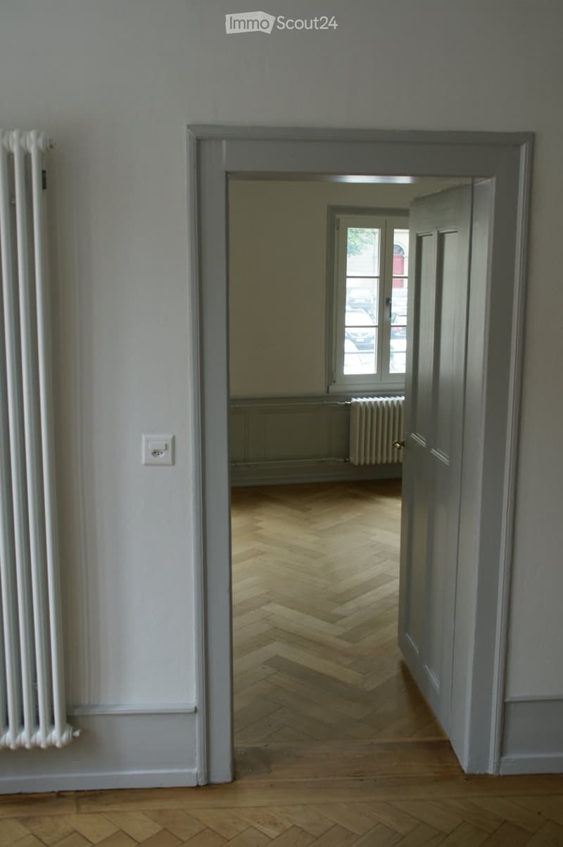 Empty hallway with wooden herringbone flooring, white walls, and a radiator on the left side. An open door leads to another room with a window and additional radiator.