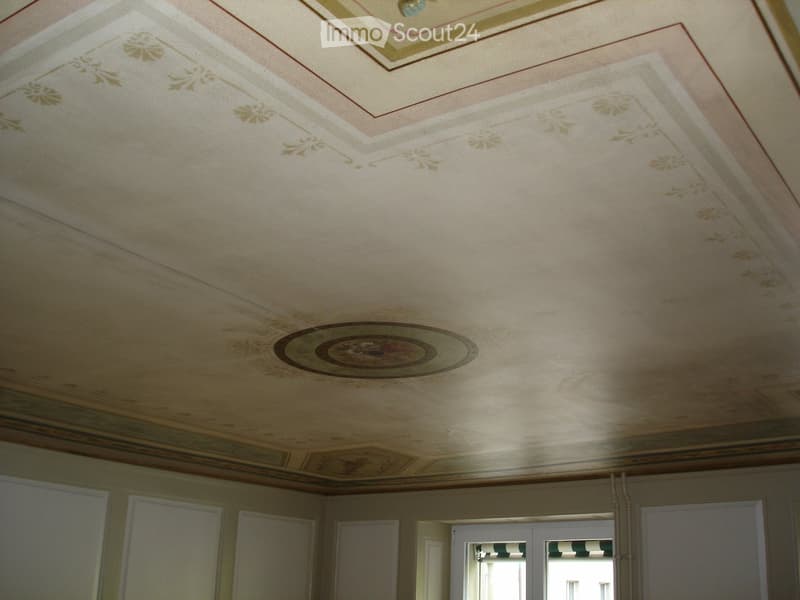 An unfurnished room with a decorative ceiling featuring floral patterns and a central round medallion. The walls are painted in a light color, and the room has wooden beams and white doors leading to other areas.