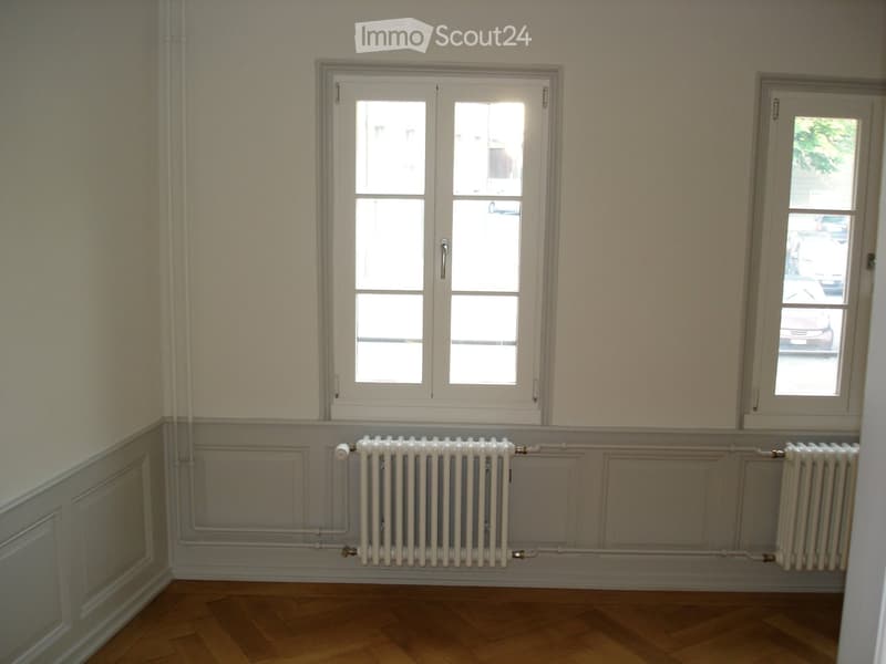Empty room with wooden flooring, white walls, two windows, and radiators below the windows.