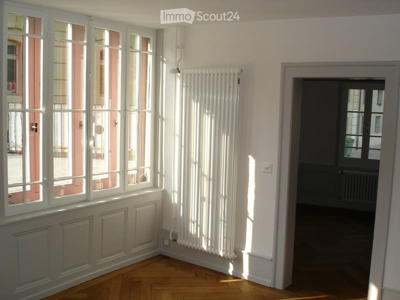 Empty room with wooden flooring, white walls, large windows, radiator, and a doorway leading to another space.