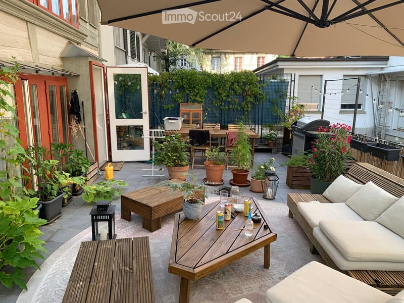 A cozy outdoor patio with a wooden sofa, coffee table, potted plants, and a large umbrella providing shade. The space includes a dining area with a table and chairs, surrounded by greenery and a view of neighboring buildings.
