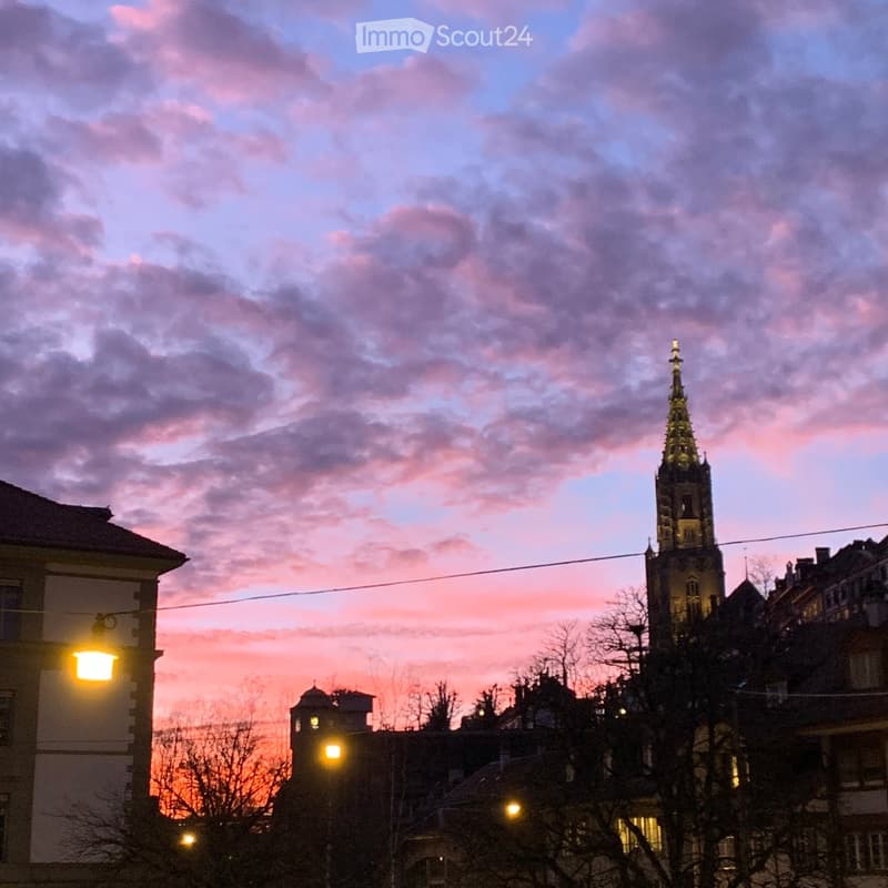 A cityscape at sunset with a prominent church tower and surrounding buildings, illuminated streetlights, and a colorful sky.