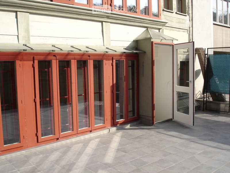 Balcony with large red-framed glass doors, tiled floor, and adjacent building entrance.