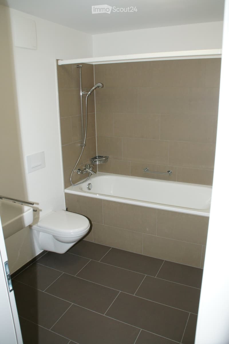 Modern bathroom with a bathtub, toilet, showerhead, and a small shower rack. The walls are tiled in beige and white, and the floor is dark brown tiles.