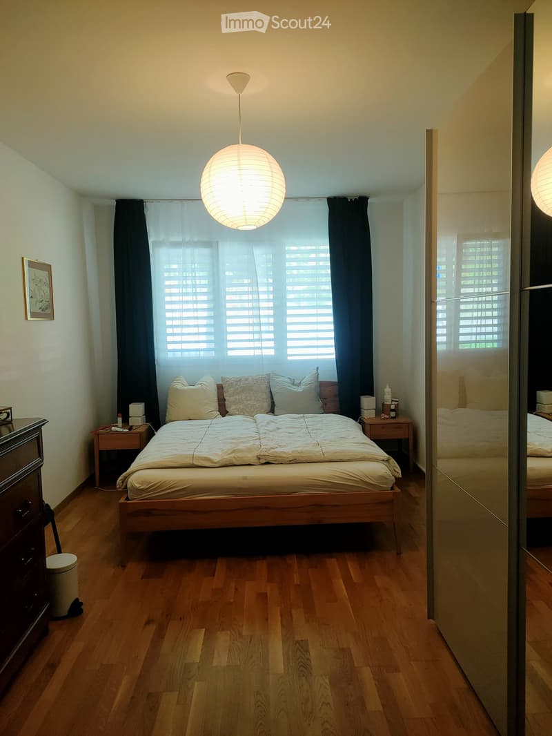 A well-lit bedroom with wooden flooring, a large bed with pillows and blankets, nightstands, a mirror, and a hanging lamp.