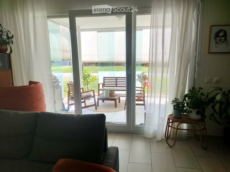 Bright living room with large sliding glass doors leading to a patio, featuring a grey sofa, a round wooden stool, a small table with potted plants, and a bench on the patio.