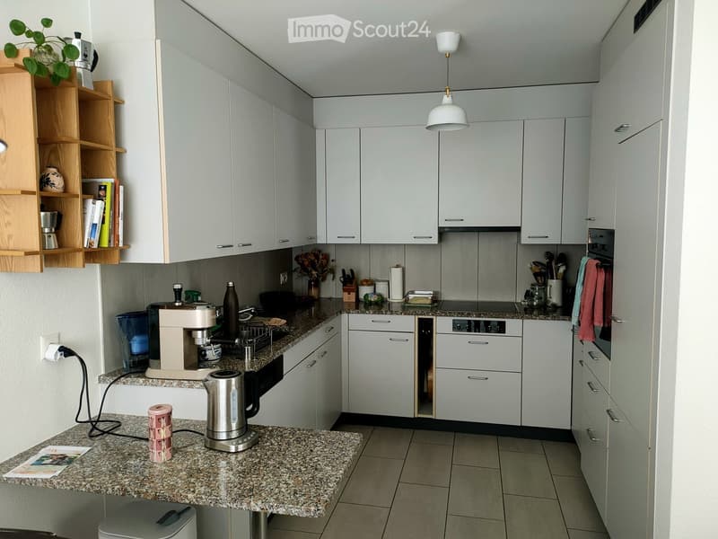 Modern kitchen with white cabinets, granite countertops, and various appliances including a coffee maker, electric kettle, and dishwasher. Wooden shelf with books and decorative items is visible on the left.