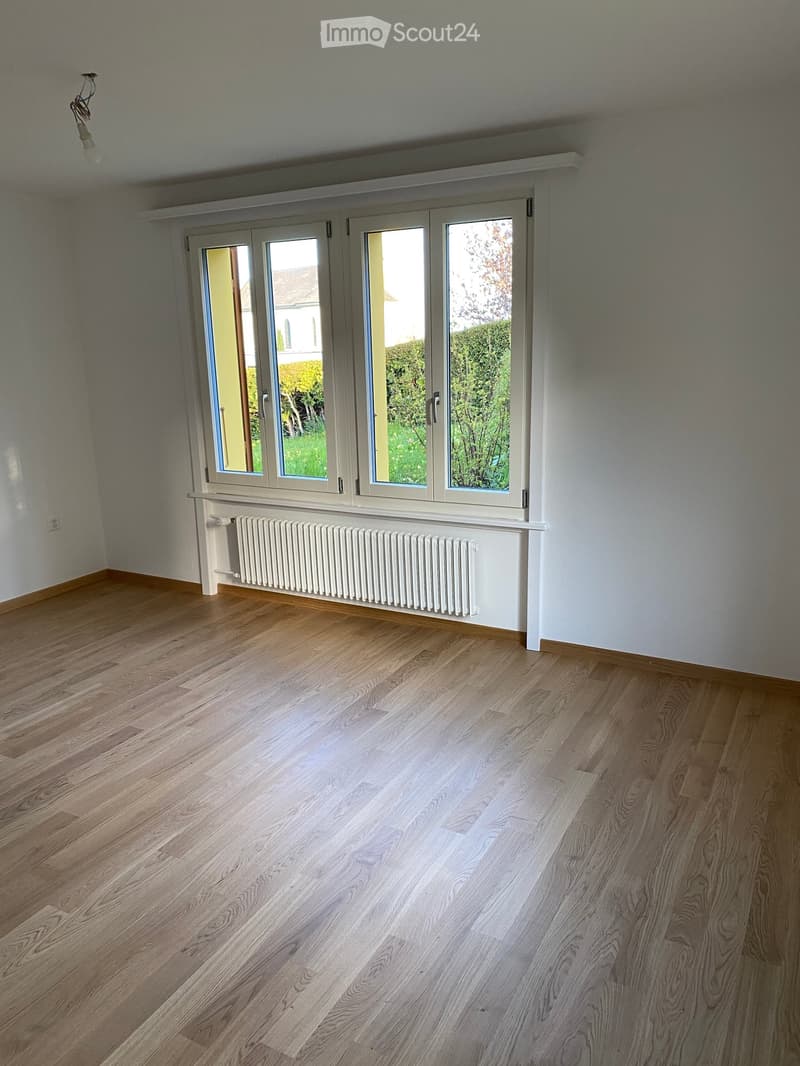 Empty room with light wooden flooring, white walls, large windows with white frames, and a white radiator below the windows.