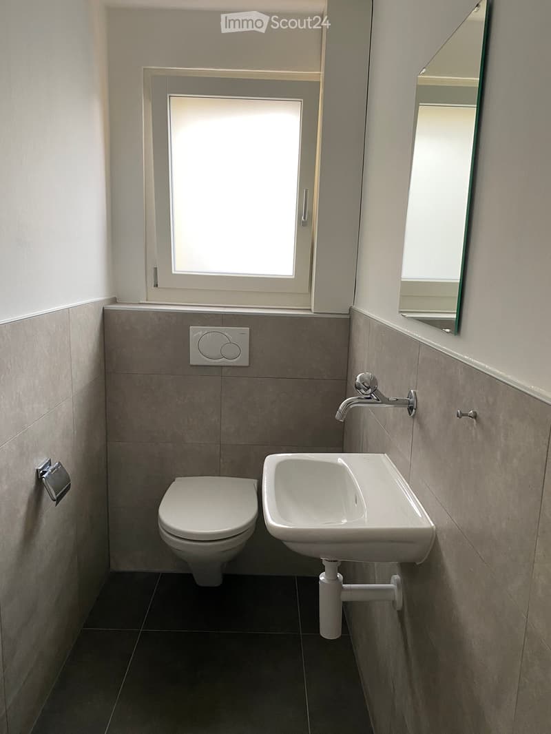 Modern bathroom with a toilet, sink, and mirror. The walls are tiled in a neutral color, and the floor is dark. A small window provides natural light.