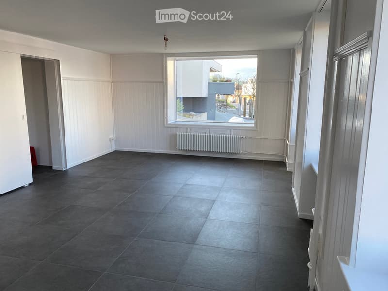 Empty room with grey tiled floor, white paneled walls, large window, radiator, and a door leading to another space.