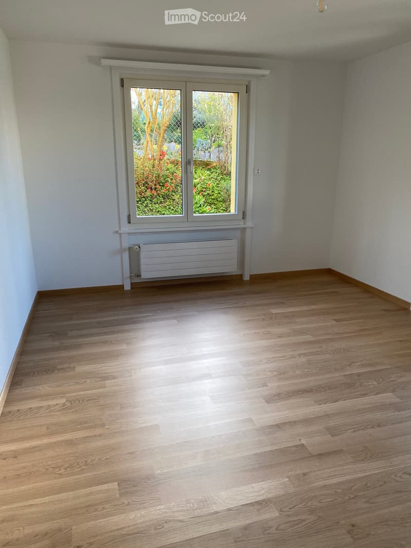 Empty room with wooden flooring, white walls, and a large window with a view of a garden. A radiator is visible below the window.