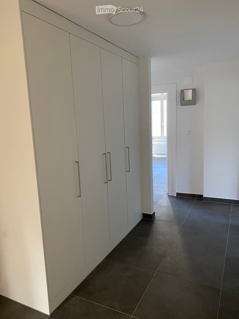 Empty hallway with white walls, dark tiled floor, built-in white cabinets with silver handles, and a recessed ceiling light. A doorway leads to another room with a window and radiator visible.