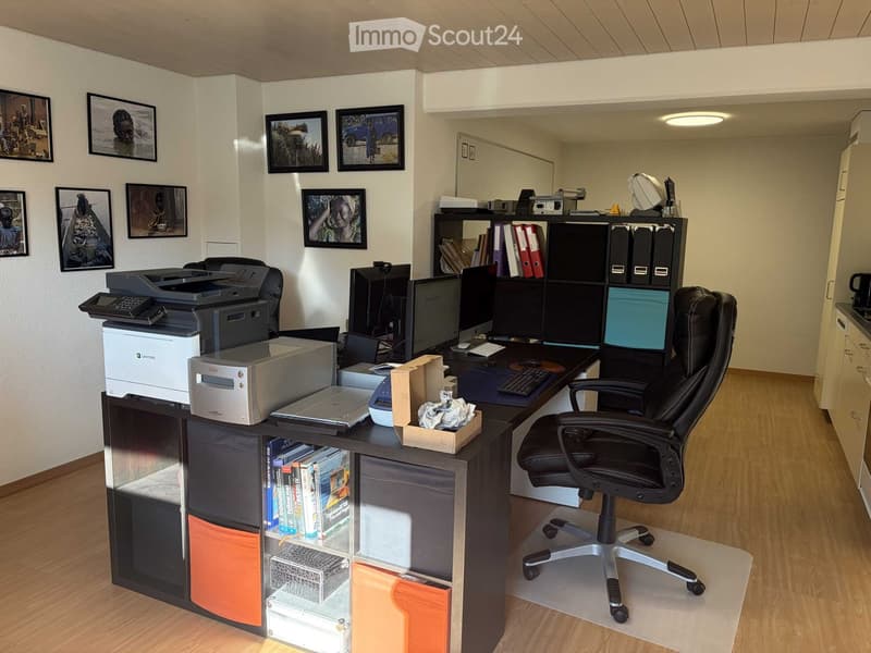 Office space with a large wooden desk, ergonomic chair, multiple computer monitors, printer, and framed artwork on the walls.