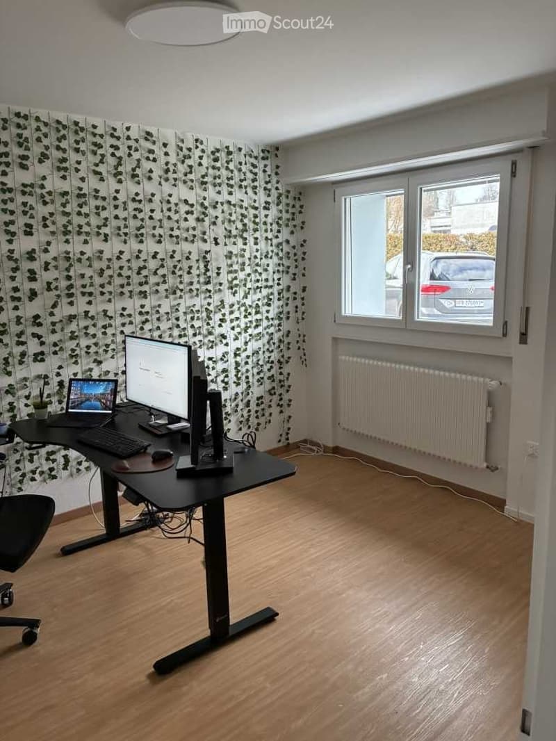 A modern, well-lit room with wooden flooring, a large window, and a black standing desk with a laptop, monitor, keyboard, and mouse. A black office chair is positioned in front of the desk.