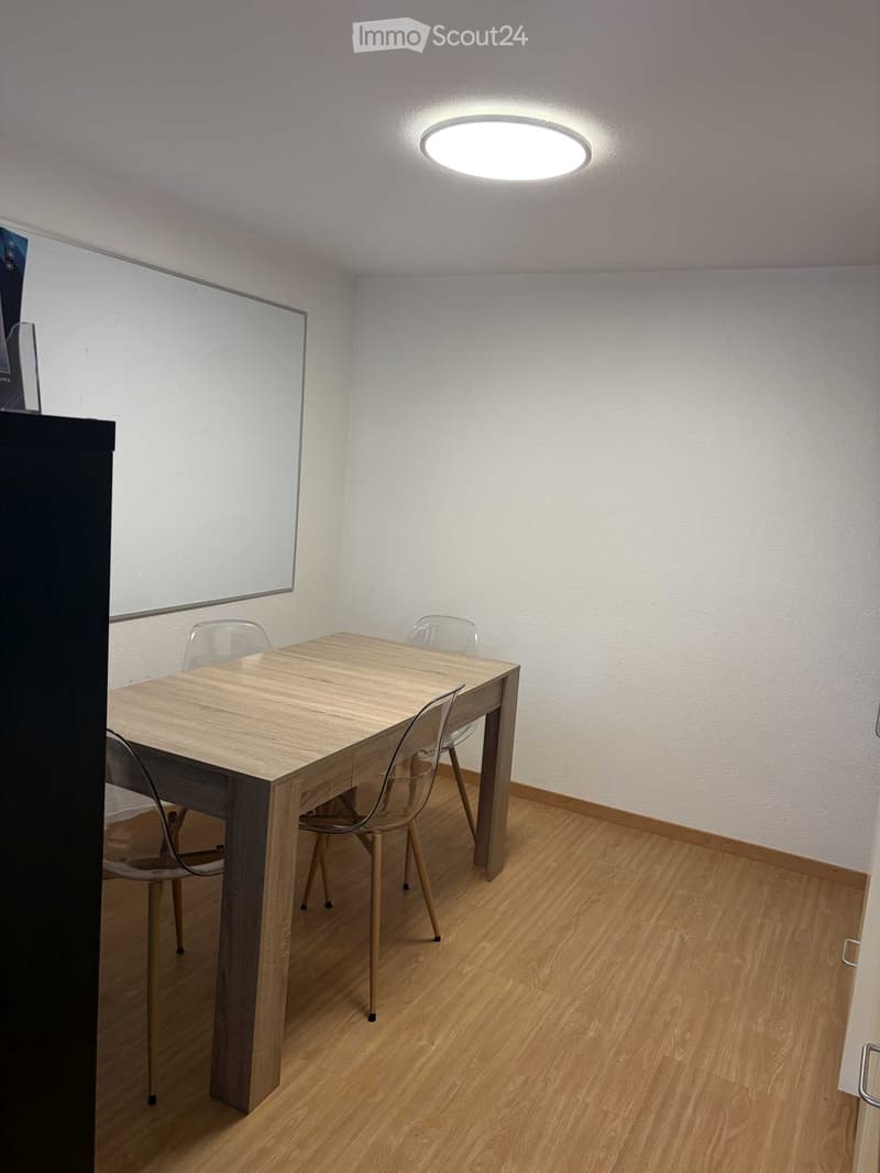 A small, well-lit room with a wooden table and four transparent chairs, a whiteboard on the wall, and a round ceiling light.