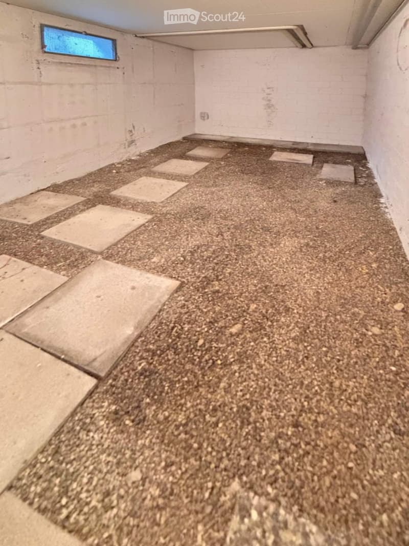 Empty room with a gravel floor and scattered square tiles, white walls, and a small window with a blue screen.